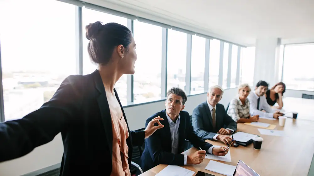 © Jacob Lund / Fotolia Eine Frau in Bürokleidung spricht zu 5 Zuhörern an einem Meetingtisch.